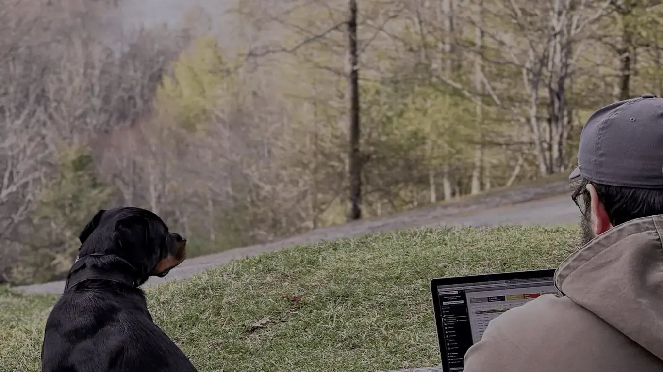 Matthew uses QuickBooks desktop on his laptop out in the woods of North Carolina while his dog sits comfortably beside him.